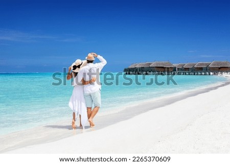 Happy couple in white clothing and with hats walks down a tropical beach with turquoise sea in the Maldives islands Royalty-Free Stock Photo #2265370609