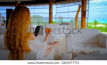 Woman with a mobile phone and a cocktail. Happy female on Maldives. A girl taking photo of a drink. Blue turquoise ocean on the background. Girl enjoys her tropical holidays. Summer travel vacation co