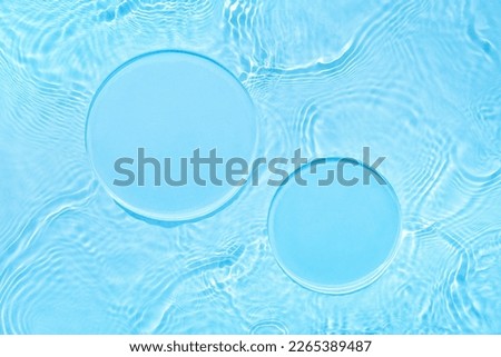 Two empty clear glass circle podiums on light blue transparent calm water texture with waves in sunlight. Abstract nature background for product presentation. Flat lay cosmetic mockup, copy space. Royalty-Free Stock Photo #2265389487