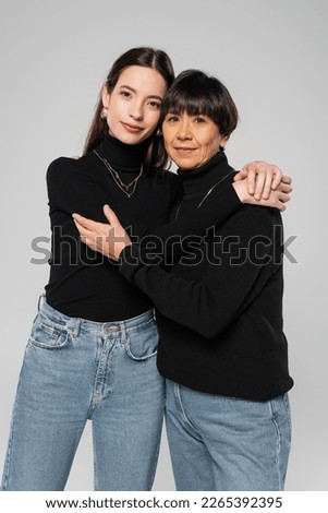 asian mother and daughter in black turtlenecks and jeans embracing and looking at camera isolated on grey Royalty-Free Stock Photo #2265392395