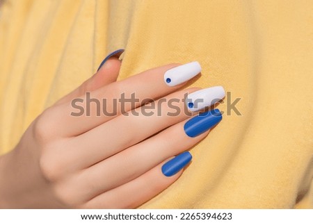 Female hand with rhinestones nail design. Glitter white and blue nail polish manicure with rhinestones nail art. Female model hand with perfect manicure and nail art on yellow fabric background. Royalty-Free Stock Photo #2265394623