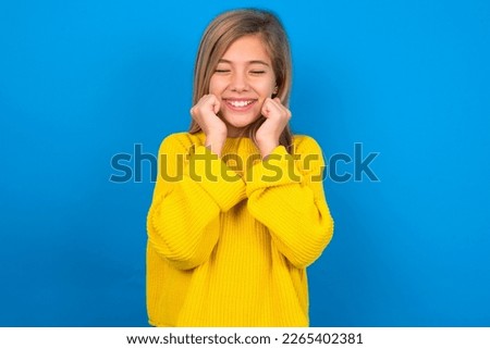beautiful caucasian teen girl wearing yellow sweater over blue wall grins joyfully, imagines something pleasant, copy space. Pleasant emotions concept. Royalty-Free Stock Photo #2265402381