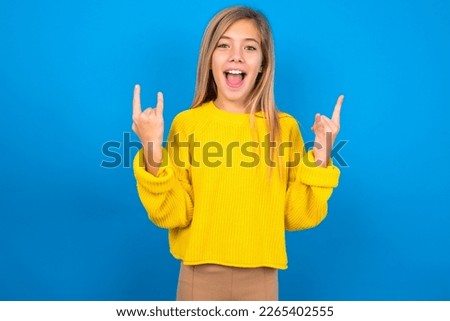 beautiful caucasian teen girl wearing yellow sweater over blue studio background makes rock n roll sign looks self confident and cheerful enjoys cool music at party. Body language concept.