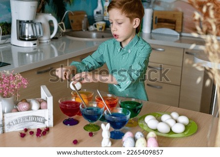 A beautiful sweet and funny red -haired boy paints eggs in the kitchen for Easter. Colored eggs, the child paints at home, ortrait.
