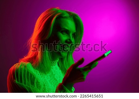 Happy smiling young woman talking on mobile phone, recording voice messages on magenta studio background in green neon light. Concept of emotions, facial expression, lifestyle, inspiration, sales, ad