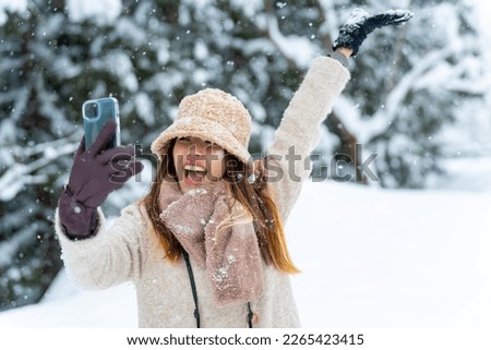 Asian woman in winter coat using mobile phone taking selfie during travel small town covered in snow. Attractive girl walking local village near forest mountain in Japan on winter holiday vacation.