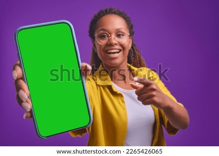 Young happy beautiful African American woman demonstrates phone with green display admiring new online shopping website or interesting news article stands in purple studio. Smartphone, applications Royalty-Free Stock Photo #2265426065