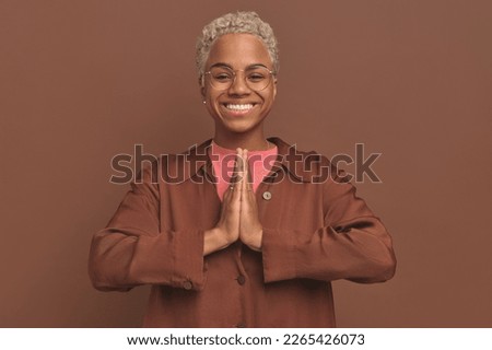 Young positive African American woman hands clasped makes prayer gesture and looks at camera with smile to ask for favor dressed in brown oversize shirt stands on studio background