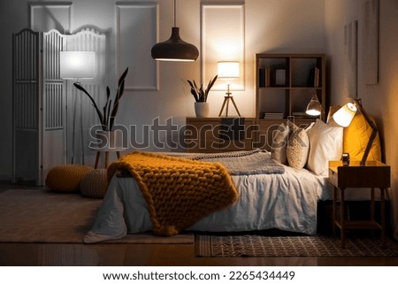 Interior of bedroom with knitted plaid on bed and glowing lamps at night Royalty-Free Stock Photo #2265434449
