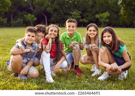 Group portrait of happy children in park. Happy school friends play in nature and enjoy summer together. Six cheerful healthy little Caucasian kids sitting on green lawn, looking at camera and smiling