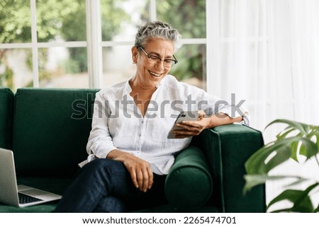 Elderly woman sitting comfortably on a couch, video calling her friends and family using a mobile phone. Retired senior woman relaxing and keeping herself engaged with technology at home. Royalty-Free Stock Photo #2265474701