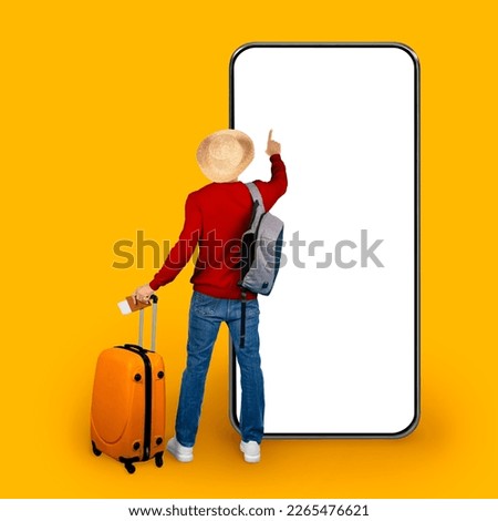 Back view of man tourist wearing summer hat, carrying backpack and orange luggage using huge smartphone with white blank screen over yellow background, booking hotel or tickets online, mockup Royalty-Free Stock Photo #2265476621