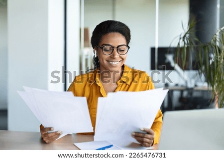 Excited black businesswoman reading papers and positive business report, sitting at workplace in modern office interior. Successful entrepreneurship career, accounting and documentation Royalty-Free Stock Photo #2265476731