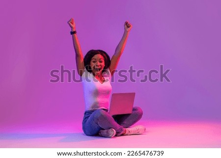 Euphoric Black Female With Laptop Celebrating Success In Neon Light, Excited Young African American Woman With Computer Sitting On Floor Over Purple Studio Background And Raising Hands, Copy Space Royalty-Free Stock Photo #2265476739