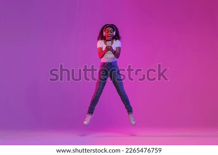 Joyful Black Woman With Smartphone In Hands Jumping In Air Over Purple Gradient Background, Positive African American Female In Headphones Messaging On Cellphone And Having Fun In Neon Light