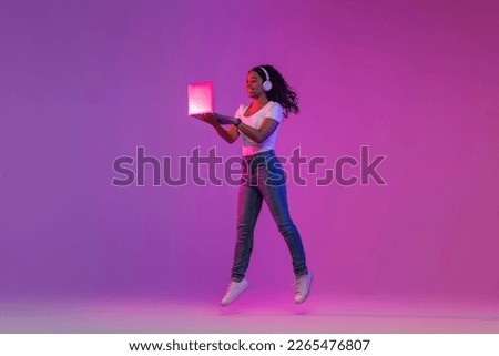Positive African American Female Jumping With Laptop In Neon Lighting, Cheerful Black Lady Wearing Wireless Headphones Using Computer For Entertainment, Enjoying Modern Technologies, Copy Space