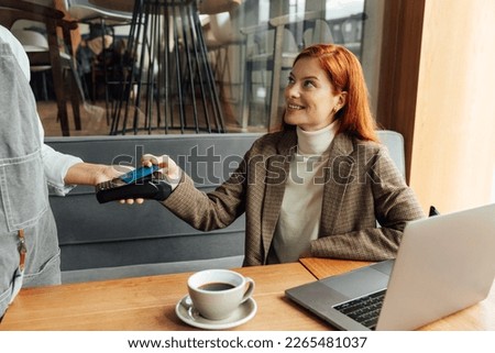 Smiling businesswoman with ginger hair paying by card in coffee shop Royalty-Free Stock Photo #2265481037