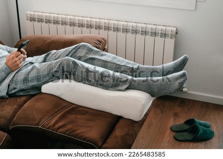 a man is leaning his legs on a leg elevation pillow, made of memory foam, resting on the brown sofa of his living room Royalty-Free Stock Photo #2265483585