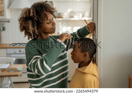 Smiling loving African American mother measuring height of child son at home, using metal ruler and pencil, marking top of head on wall, mom taking measurement while boy standing against flat surface Royalty-Free Stock Photo #2265494379