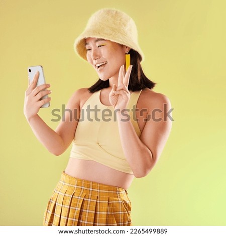 Selfie, peace sign and smile of Asian woman in studio isolated on a yellow background. Makeup, fashion hat and young female model with v hand gesture taking pictures for social media or happy memory.