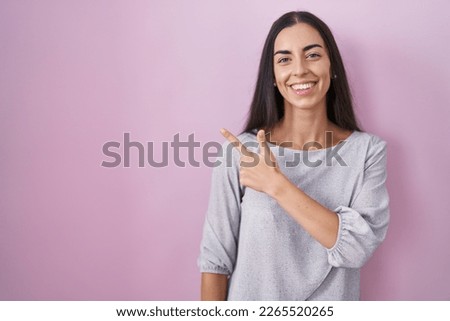 Young brunette woman standing over pink background cheerful with a smile of face pointing with hand and finger up to the side with happy and natural expression on face 