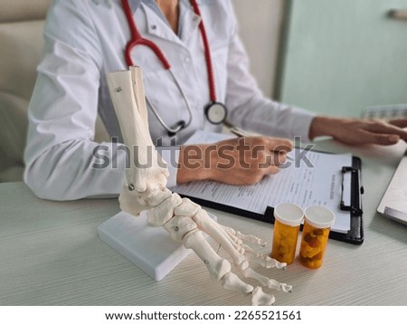Doctor traumatologist writes out prescription for pills and leg skeleton. Pain in legs with osteoarthritis of bones and treatment with calcium and protein Royalty-Free Stock Photo #2265521561