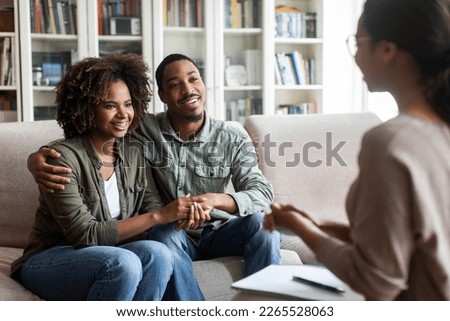 Happy beautiful young black couple in casual sitting on couch in cozy counselor office, embracing, holding hands and smiling after successful marital family therapy, copy space