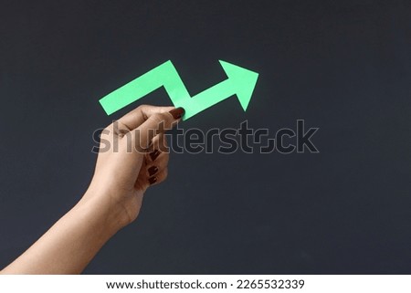 Hand hold arrow cutted from solid sheet of green paper and curved up of one side showing growth of stock market or up direction isolated on dark background with copy space.  Royalty-Free Stock Photo #2265532339