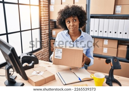 Black woman with curly hair working at small business ecommerce holding box smiling looking to the side and staring away thinking. 