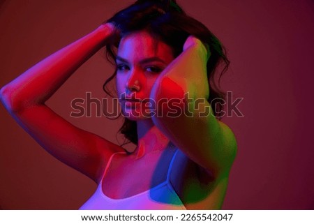 Closeup portrait of young beautiful girl with long dark hair posing over dark red background in neon light. Concept of beauty, skin care, cosmetology, wellness, makeup. Model with well-kept skin