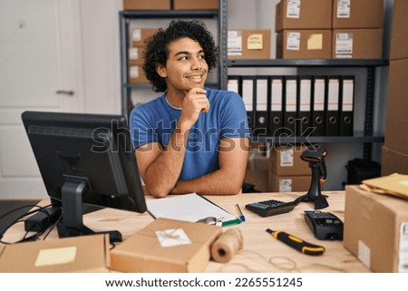 Hispanic man with curly hair working at small business ecommerce with hand on chin thinking about question, pensive expression. smiling and thoughtful face. doubt concept. 