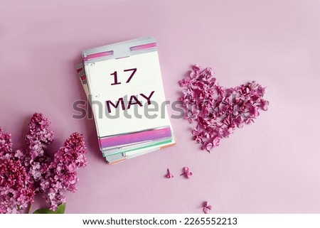 Calendar for May 17: a desk calendar with the numbers 17, the name of the month of May in English, a branch of lilac, a heart of lilac inflorescences on a pastel background, top view