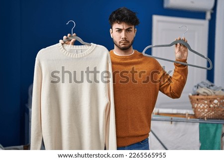 Hispanic man with beard holding sweater on hanger at laundry room skeptic and nervous, frowning upset because of problem. negative person. 