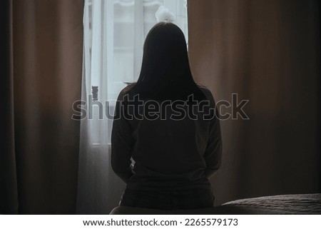 Silhouette of a young woman in pajamas against the background of a window at home on a bed with seasonal affective disorder or depression. The concept of winter depression due to lack of sunlight Royalty-Free Stock Photo #2265579173