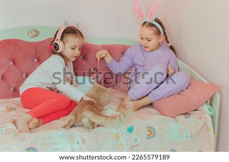 two sister girls play on the bed with a red domestic cat, listen to music on headphones, the older girl has a bandage with rabbit ears on her head, family preparation for Easter