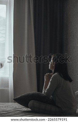 A sad young woman in pajamas with seasonal affective disorder or depression sits alone on the bed and looks out the window. The concept of winter depression due to lack of sunlight Royalty-Free Stock Photo #2265579191