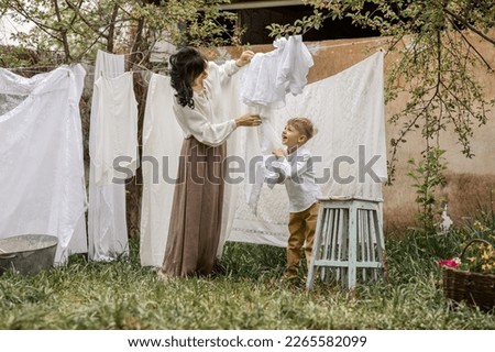 a blond boy of primary school age in a white shirt helps his mother hang freshly laundered laundry on ropes in the spring in the garden of a country house. the concept of village life, healthy lifesty Royalty-Free Stock Photo #2265582099