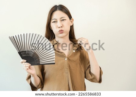 Suffering summer heat stroke, hot weather, tired asian young woman, girl sweaty and thirsty, refreshing with hand in blowing, wave fan to ventilation when temperature high at home, house on background Royalty-Free Stock Photo #2265586833