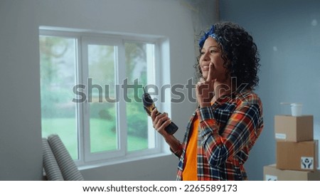 Young African American woman with a cordless screwdriver in hand. Black female thinking something against background of an apartment in process of repair. Concept of apartment renovation.