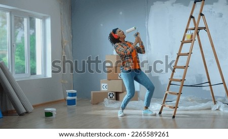 Young african american woman in big wireless red headphones is enjoying music. Black female in checkered shirt sings into paint roller as if into microphone and dances. Ladder, cardboard boxes, window Royalty-Free Stock Photo #2265589261