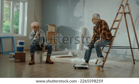 An upset elderly man with a roller in his hands sits on the stairs. An elderly woman sitting on a large can of paint and looks tiredly at a man.