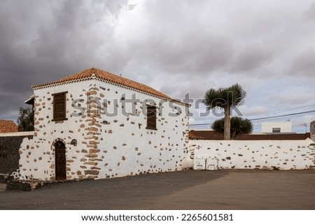 Very traditional building with a white facade, green details, stones in the facade and orange roof. Empty yard around. Cloudy sky. Antigua, Fuerteventura, Canary Islands, Spain.