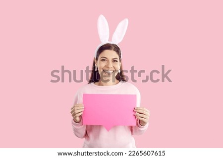 Portrait of cheerful woman with bunny ears in pink sweatshier holding pink rectangular paper word bubble or speech balloon on pink background. Celebrating easter, spring holidays, message concept.