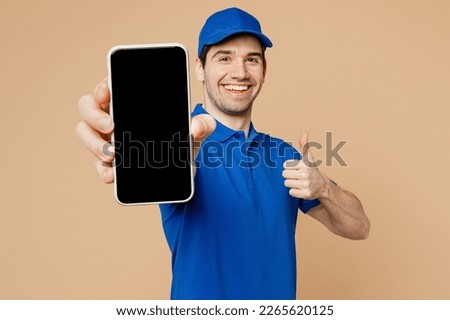 Delivery guy employee man wearing blue cap t-shirt uniform workwear work as dealer courier hold in hand use mobile cell phone with blank screen area show thumb up isolated on plain beige background Royalty-Free Stock Photo #2265620125