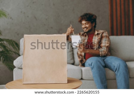 Young Indian man wear casual clothes eating Chinese food cuisine and takeaway carton container box sit on grey sofa couch stay at home hotel flat rest relax spend free spare time in living room indoor