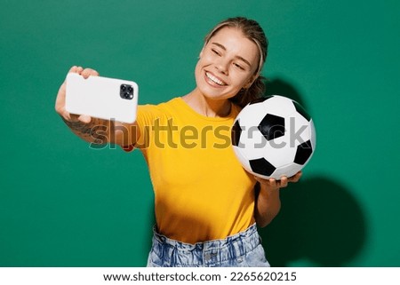 Young fun woman fan wear basic yellow t-shirt cheer up support football sport team hold in hand soccer ball watch tv live stream do selfie shot on mobile cell phone isolated on dark green background
