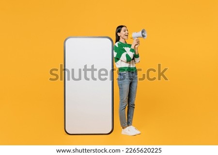 Full body young latin woman wear casual cozy green knitted sweater big huge blank screen mobile cell phone smartphone with area scream in megaphone isolated on plain yellow background studio portrait