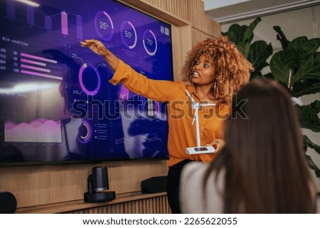 An African American woman is in a boardroom presenting a renewable energy strategy plan and pointing on research displayed on the screen and holding a windmill model in her hands.