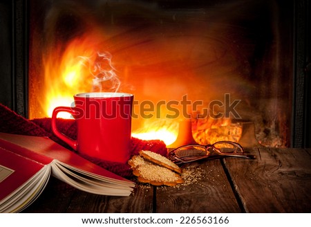 Hot tea or coffee in a red mug, ginger cookies, book and glasses on vintage wood table. Fireplace as background. Christmas or winter warming drink. Layout with free text space.