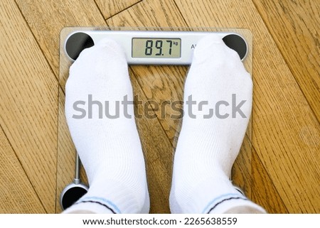 A man in white socks weighing himself on a transparent electronic floor scale, the weight reading is 89 kg, top view